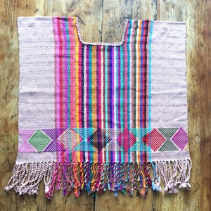 Op de afbeelding: Een geweven poncho met een lichtroze basis en verticale strepen in tinten roze, oranje, geel, groen, blauw en paars. Het onderste gedeelte toont een rij ruitvormige patronen in levendige kleuren, afgewerkt met lange gedraaide franjes.