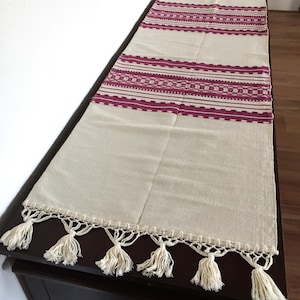 Peut inclure: Chemin de table crème avec des motifs géométriques bordeaux et des glands décoratifs. Le chemin de table présente un motif tissé avec trois bandes de détails à motifs. Les glands sont blancs et pendent du bord inférieur.