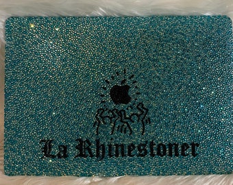May include: A rectangular laptop cover fully encrusted with shimmering teal rhinestones. The center features a black rhinestone Apple logo above a black rhinestone graphic of dancing figures and the text La Rhinestoner in black.