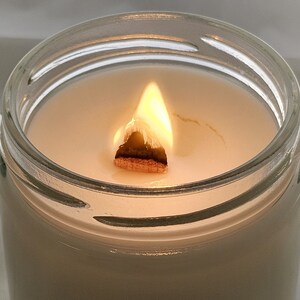 Puede incluir: Una vela blanca en un tarro de vidrio transparente con una mecha de madera que se quema. La llama es de color naranja y amarillo brillante.