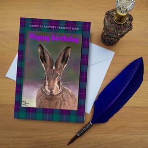 Peut inclure: Carte d'anniversaire avec une illustration de lièvre, bordure écossaise et texte rose « Happy birthday ». Une plume bleue, un flacon décoratif et une enveloppe sont disposés sur une surface en bois, créant une scène festive.