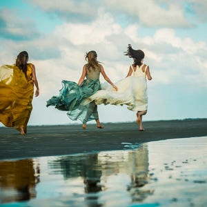May include: Three women in flowing dresses run along a sandy beach with the ocean in the background. The dresses are yellow, green, and white.