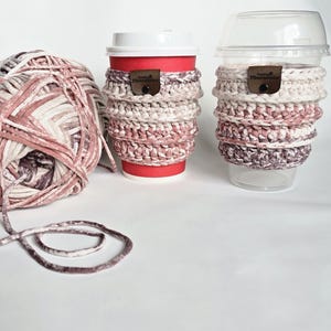 May include: Two coffee cups with crocheted cozies in shades of pink, cream, and brown. A ball of yarn in matching colors sits to the left. The cozies have a leather tag that reads "Good Vibes Fiberations".