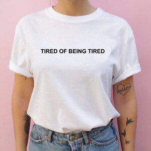 May include: White t-shirt with the text "TIRED OF BEING TIRED" printed in black on the front.
