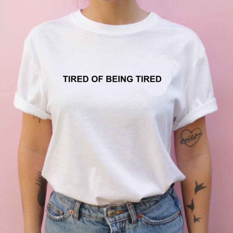 May include: White t-shirt with the text "TIRED OF BEING TIRED" printed in black on the front.