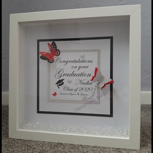 May include: A white frame with a black and white background. The background has a silver glitter overlay. The text reads "Congratulations on your Graduation Nadia Class of 2020 Love from Lizana &  Jayden... Handmade Gifts". There are red and black paper butterflies and a graduation cap.