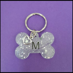 May include: A bone-shaped pet tag in light purple with silver glitter and the letter "M". It has a small silver heart charm and a silver keyring. The tag is designed for pets.