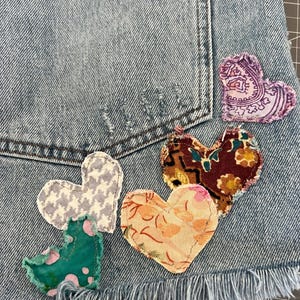 May include: Close-up of a light blue denim garment, possibly shorts, adorned with five heart-shaped fabric patches. The patches feature various patterns and colors, including paisley, floral, and houndstooth. The garment has a frayed hem.