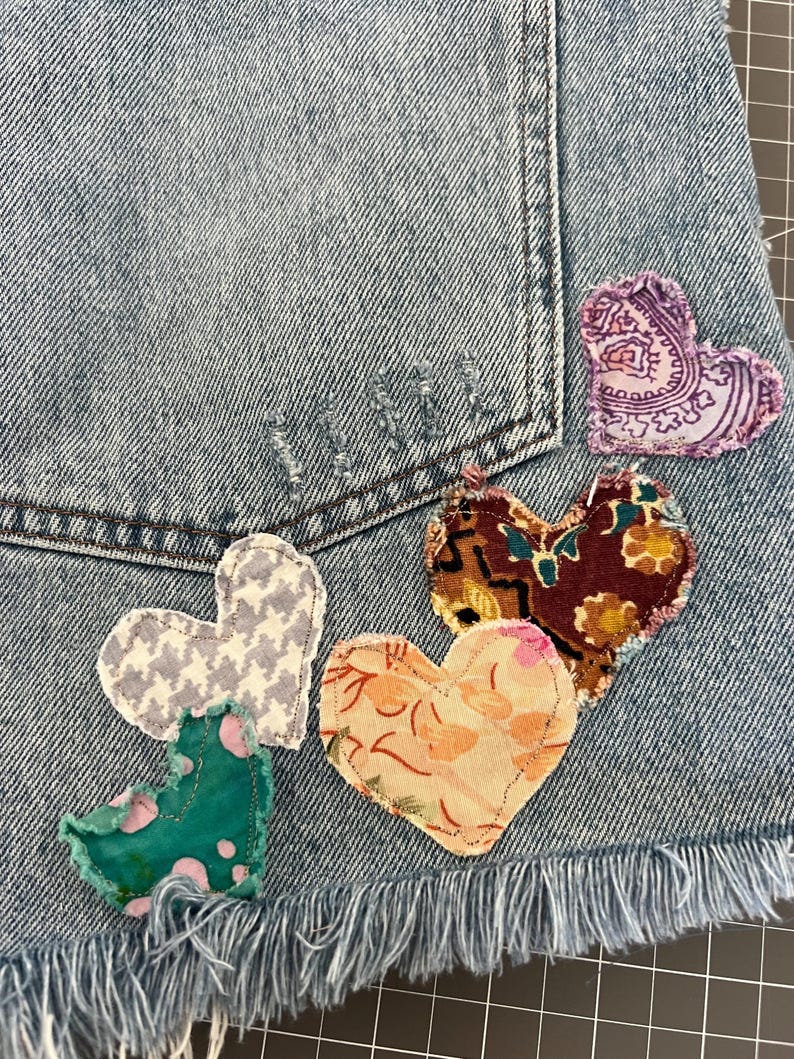May include: Close-up of a light blue denim garment, possibly shorts, adorned with five heart-shaped fabric patches. The patches feature various patterns and colors, including paisley, floral, and houndstooth. The garment has a frayed hem.