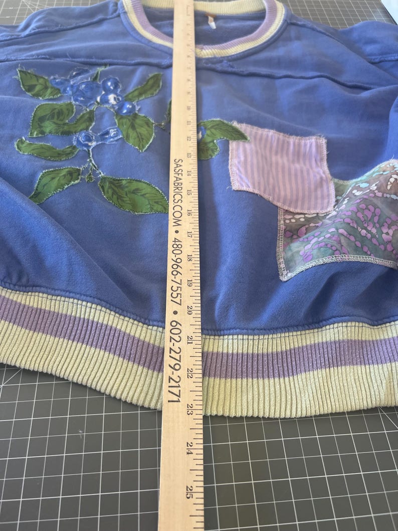 Puede incluir: Una sudadera azul con cuello redondo y dobladillo acanalado. La sudadera presenta un dise&ntilde;o floral con bayas azules y hojas verdes. Tiene un bolsillo y ribete de color lila claro. La longitud de la sudadera es de aproximadamente 63.5 cm.