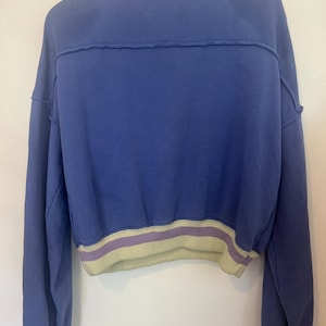 Puede incluir: Una sudadera corta azul con cuello y dobladillo de canal&eacute;. El cuello presenta una raya verde claro y morada, y el dobladillo y los pu&ntilde;os tienen una raya a juego. La sudadera tiene mangas largas y un corte holgado.