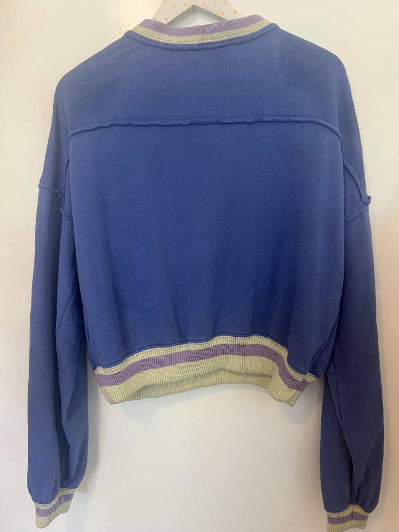 Puede incluir: Una sudadera corta azul con cuello y dobladillo de canal&eacute;. El cuello presenta una raya verde claro y morada, y el dobladillo y los pu&ntilde;os tienen una raya a juego. La sudadera tiene mangas largas y un corte holgado.