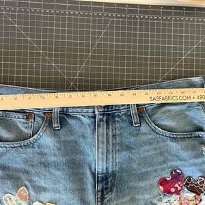 May include: Light blue denim shorts with decorative patches, including heart and floral designs. A wooden ruler is placed across the top of the shorts. The shorts have a distressed look with visible wear and tear.