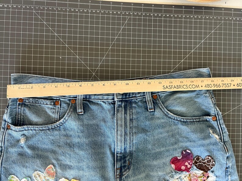May include: Light blue denim shorts with decorative patches, including heart and floral designs. A wooden ruler is placed across the top of the shorts. The shorts have a distressed look with visible wear and tear.