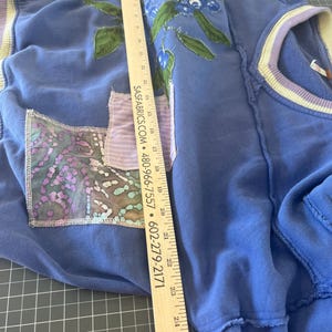 Puede incluir: Una sudadera azul con un dise&ntilde;o floral y un bolsillo de parche. El dise&ntilde;o floral presenta flores azules y hojas verdes. La sudadera tiene un aspecto desgastado con bordes deshilachados. Una regla est&aacute; colocada sobre la sudadera.