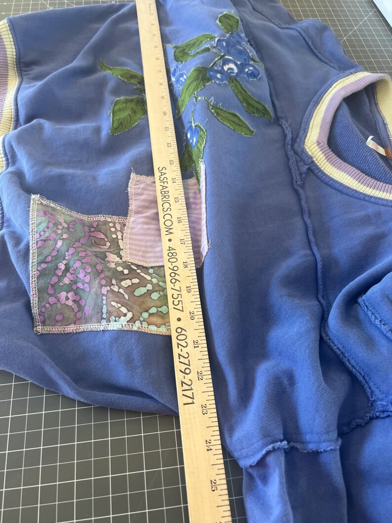 Puede incluir: Una sudadera azul con un dise&ntilde;o floral y un bolsillo de parche. El dise&ntilde;o floral presenta flores azules y hojas verdes. La sudadera tiene un aspecto desgastado con bordes deshilachados. Una regla est&aacute; colocada sobre la sudadera.