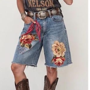 May include: A charcoal gray short-sleeve t-shirt with Willie Nelson graphic text, blue denim Bermuda shorts featuring floral embroidery and a frayed hem, a brown leather belt with a silver buckle, and brown leather cowboy boots with stitching.