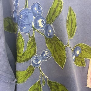 Puede incluir: Una sudadera azul claro con un dise&ntilde;o bordado de ar&aacute;ndanos y hojas verdes. El dise&ntilde;o floral est&aacute; cosido a la tela con un borde deshilachado. La sudadera tiene un cuello y mangas desgastados.