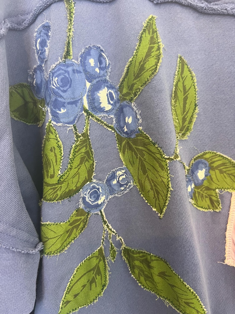 Puede incluir: Una sudadera azul claro con un dise&ntilde;o bordado de ar&aacute;ndanos y hojas verdes. El dise&ntilde;o floral est&aacute; cosido a la tela con un borde deshilachado. La sudadera tiene un cuello y mangas desgastados.