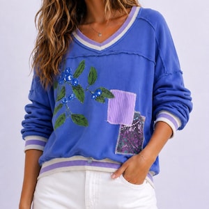Puede incluir: Una sudadera azul con cuello en V con un dise&ntilde;o de ar&aacute;ndanos y hojas. La sudadera tiene un ribete a rayas blancas y lavanda en el cuello, los pu&ntilde;os y el dobladillo. Parches de tela cosidos en la parte delantera.