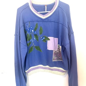 Puede incluir: Una sudadera azul de manga larga con cuello en V y dobladillo corto. La sudadera presenta un dise&ntilde;o de ar&aacute;ndanos bordados y dos parches en la parte delantera. El cuello y el dobladillo tienen detalles a rayas.