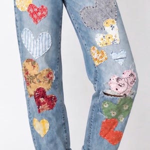 May include: Light blue denim jeans featuring a variety of heart-shaped fabric patches in assorted colors and patterns, including floral, striped, and solid designs. The jeans have a relaxed fit with rolled cuffs at the bottom of each leg.
