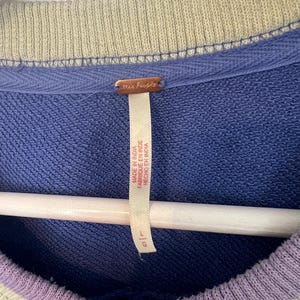 Puede incluir: Primer plano de una sudadera azul con cuello y pu&ntilde;os a rayas verdes y moradas. La etiqueta dice "free people" y "Made in India". La sudadera est&aacute; colgada en una percha blanca.
