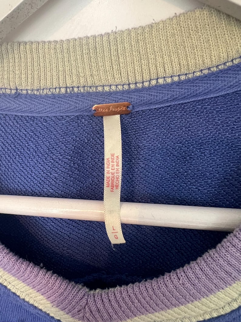 Puede incluir: Primer plano de una sudadera azul con cuello y pu&ntilde;os a rayas verdes y moradas. La etiqueta dice "free people" y "Made in India". La sudadera est&aacute; colgada en una percha blanca.