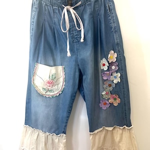 May include: Blue denim culottes with a drawstring waist and decorative floral appliques. The pants feature a cream-colored lace trim at the hem and a floral embroidered pocket. The style is casual and bohemian.