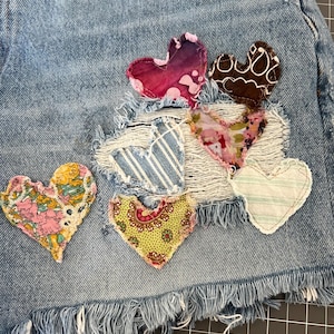 May include: Close-up of denim shorts with frayed edges, adorned with colorful, fabric heart patches. The hearts feature various patterns and textures, including floral, striped, and paisley designs. The shorts are a light blue wash.