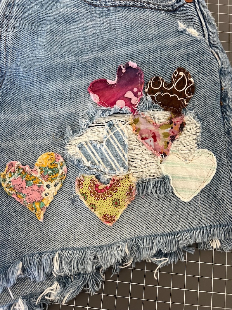 May include: Close-up of denim shorts with frayed edges, adorned with colorful, fabric heart patches. The hearts feature various patterns and textures, including floral, striped, and paisley designs. The shorts are a light blue wash.