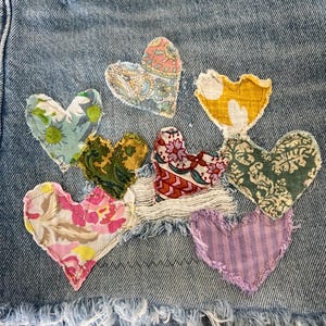 May include: Close-up of denim shorts with frayed edges, adorned with appliqued fabric hearts in various patterns and colors, including floral, paisley, and striped designs. The hearts are stitched onto the denim.
