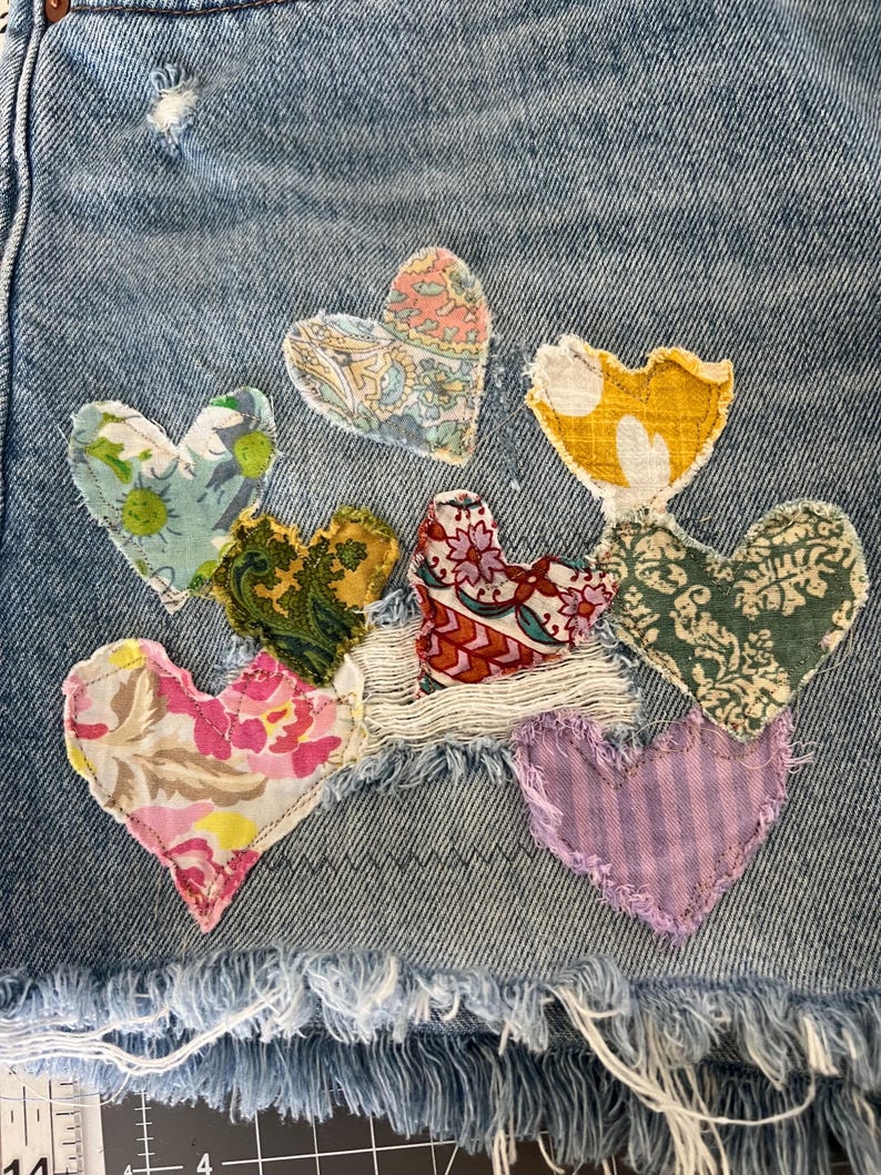 May include: Close-up of denim shorts with frayed edges, adorned with appliqued fabric hearts in various patterns and colors, including floral, paisley, and striped designs. The hearts are stitched onto the denim.