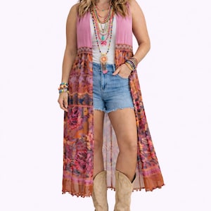 May include: A woman wearing a layered necklace, bracelets, denim shorts, and a long floral vest over a white top and pink vest. She is also wearing beige cowboy boots. The vest features a floral pattern in shades of pink, orange, and brown.