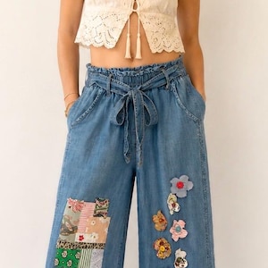 May include: A light blue denim pants with floral and patchwork embellishments. The pants have a high waist with a tie, and a ruffled hem. The top is a cream-colored, short-sleeved blouse with a lace design.