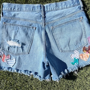 May include: Light blue denim shorts with a frayed hem and distressed details. The shorts feature patches of colorful, patterned hearts on the back pockets and sides, adding a unique, handcrafted touch to the garment.