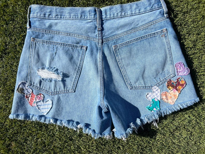 May include: Light blue denim shorts with a frayed hem and distressed details. The shorts feature patches of colorful, patterned hearts on the back pockets and sides, adding a unique, handcrafted touch to the garment.