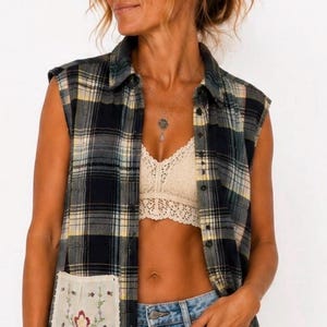 May include: A sleeveless plaid button-down shirt in navy, yellow, and white with a cream-colored lace trim along the hem. The shirt features a single cream-colored patch pocket on the front with a small floral embroidery design in red and green.