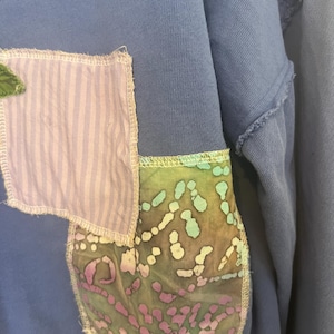 Puede incluir: Primer plano de una sudadera azul con parches decorativos. Un parche es de tela a rayas moradas claras y blancas, y el otro es de tela estampada verde, azul y morada. Los parches est&aacute;n cosidos a la sudadera con hilo blanco.