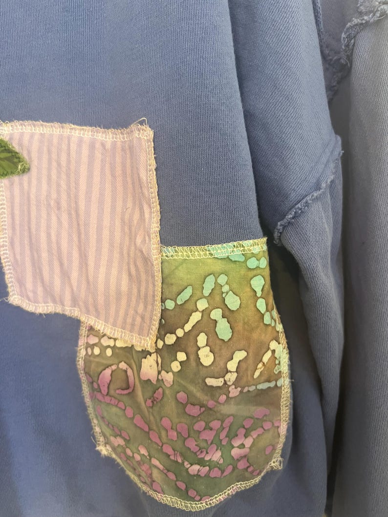 Puede incluir: Primer plano de una sudadera azul con parches decorativos. Un parche es de tela a rayas moradas claras y blancas, y el otro es de tela estampada verde, azul y morada. Los parches est&aacute;n cosidos a la sudadera con hilo blanco.
