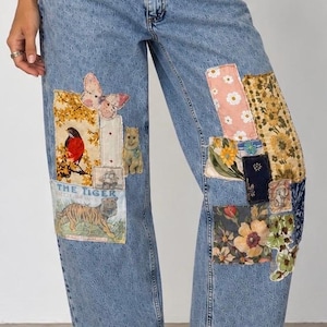May include: Light blue denim jeans featuring a subtle embossed pattern and various fabric patches. The patches include a butterfly, a bird, a tiger with the text THE TIGER, floral prints, and geometric designs. The jeans have a straight leg and raw hem.