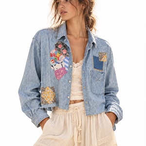 May include: A light blue denim button-down shirt featuring a cropped, raw-edge hem and various colorful floral and patterned fabric patches sewn onto the front. The shirt is worn over a cream-colored lace camisole and light-colored tiered trousers.