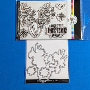 May include: Clear stamp set featuring a reindeer with antlers decorated with ornaments, snowflakes, and the text "Here's to a festive season." Also includes matching metal dies for crafting. The set is packaged on a blue background.