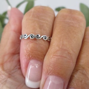 May include: A silver ring with a delicate, swirling design. The ring is worn on a finger with a French manicure.