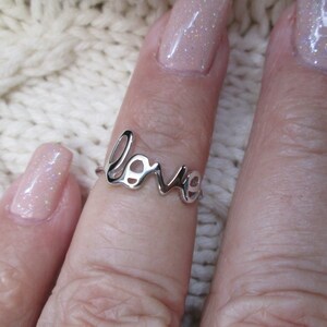 May include: A silver ring with the word "love" spelled out in a cursive font. The ring is on a finger with light pink nail polish with glitter.