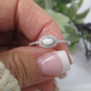 May include: A silver ring with an oval opal center stone, surrounded by a halo of small, clear gemstones. The ring band is also adorned with small, clear gemstones. The ring is held up by a hand with a white sweater in the background.