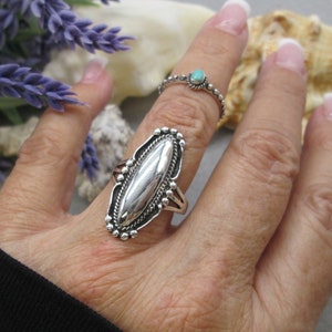 May include: A silver ring with an oval-shaped stone set in a detailed design. The ring is on a finger with a turquoise ring on another finger.
