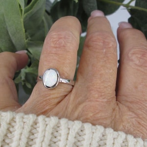 May include: A silver ring with an oval, white gemstone is displayed on a hand. The ring's band is thin and polished. The gemstone is set in a bezel setting. The hand is wearing a cream-colored sweater.