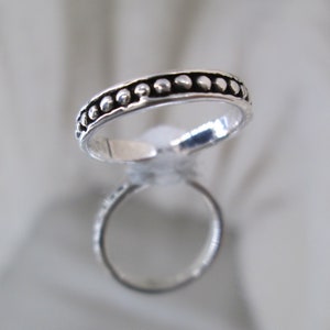 May include: A silver ring with a band of small, evenly spaced, raised dots. The ring is highly reflective, with a clear reflection visible below it. The ring is likely made of sterling silver and is a simple, elegant design.