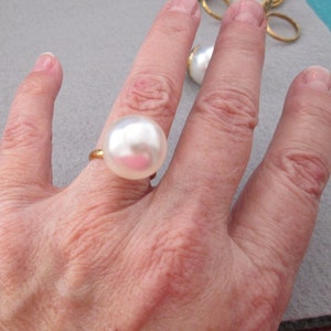 May include: A large, white faux pearl ring with a gold-colored band. The ring features a prominent, round pearl. Other rings with similar pearls are visible in the background. The ring is a statement piece of jewelry.